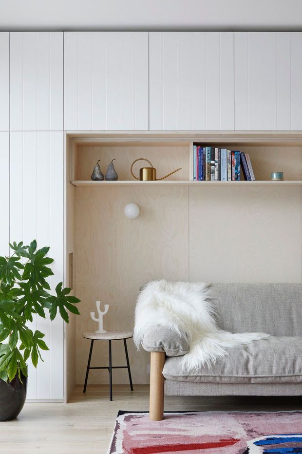 A calm, light-filled living room vignette with a grey upholstered sofa, white faux-fur throw, pale timber built-in shelving and cabinetry, books, a brass watering can, and restrained decorative objects. White vertical wall panelling, pale timber flooring, a large indoor plant, side table, and colourful patterned rug complete the softly styled interior.