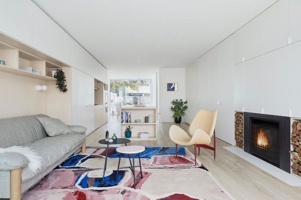 Bright, compact contemporary living room with a grey sofa, sculptural cream lounge chair, colourful abstract rug, round nesting coffee tables, pale built-in cabinetry and open shelving. A central timber-toned shelving unit partially separates the seating area from a glazed dining or outdoor connection at the rear. The room features a minimalist white fireplace wall with integrated firebox and stacked firewood niches, pale timber flooring, and abundant natural light.