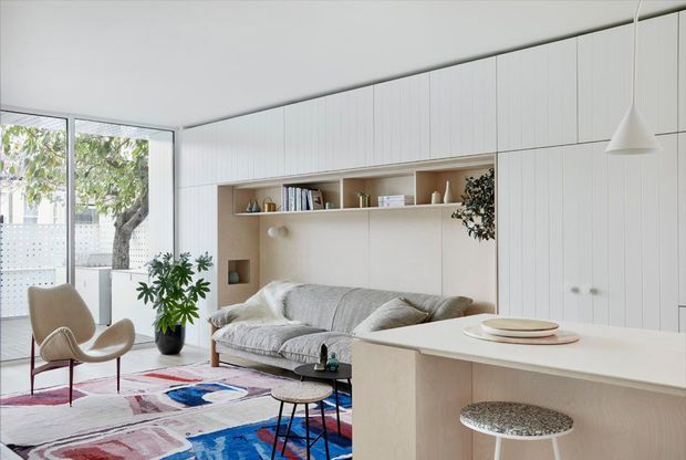 Bright, compact open-plan living area with a pale built-in wall of vertically panelled cabinetry and integrated shelving around a textured grey sofa. A light kitchen or breakfast counter occupies the foreground with two stone-look stools, while a sculptural lounge chair, colourful patterned rug, indoor plants, books and simple pendant lighting add visual interest. Full-height glazing connects the room to a leafy outdoor area. The overall design is restrained, light and contemporary with warm natural accents.