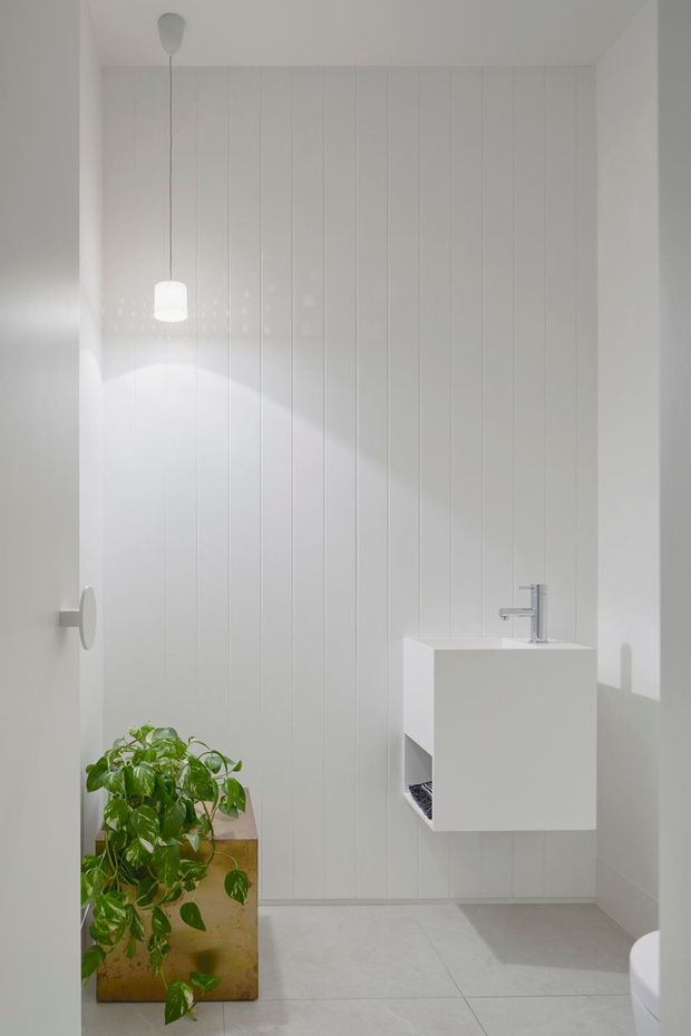 A minimalist white bathroom with vertically panelled wall cladding, pale large-format floor tiles, a wall-mounted white vanity with integrated basin and chrome tapware, and a partially visible toilet. A hanging pendant light provides a soft focal glow, while a leafy potted plant adds the only strong natural colour accent.