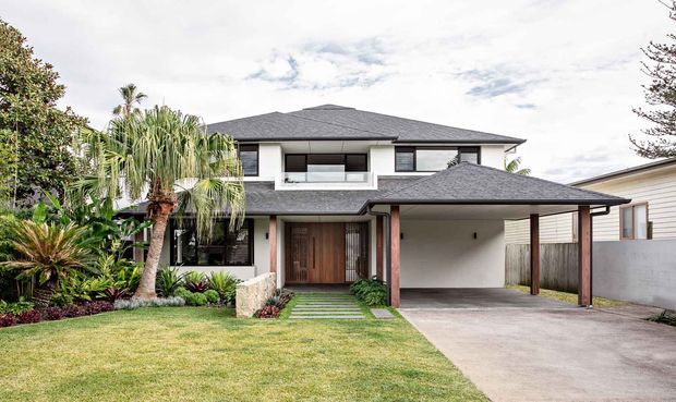 Street-facing photograph of a two-storey contemporary residence with a dark hipped roof, white rendered walls, timber front door and accents, broad upper-level glazing, landscaped front garden, and an integrated covered carport.