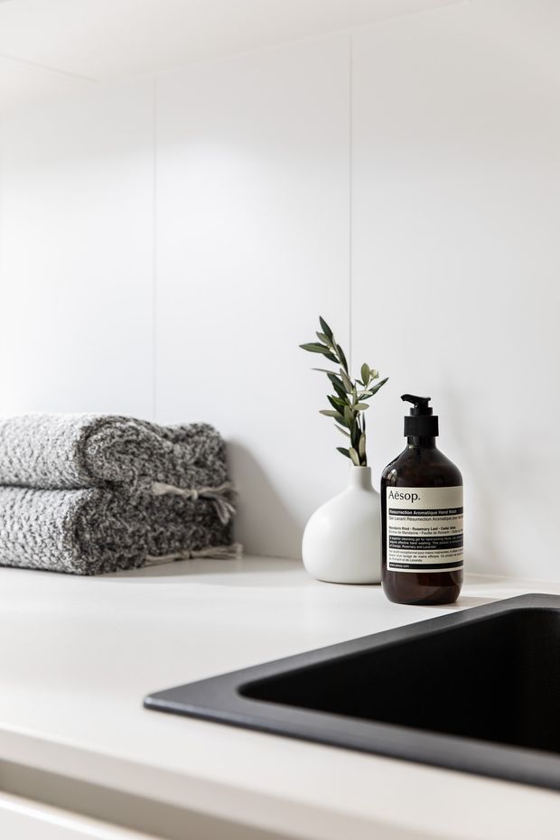 Close-up of a minimalist bathroom vanity with a matte black inset basin, white countertop and white paneled wall. Folded grey towels, a small white vase with greenery, and a dark amber soap dispenser sit on the counter.