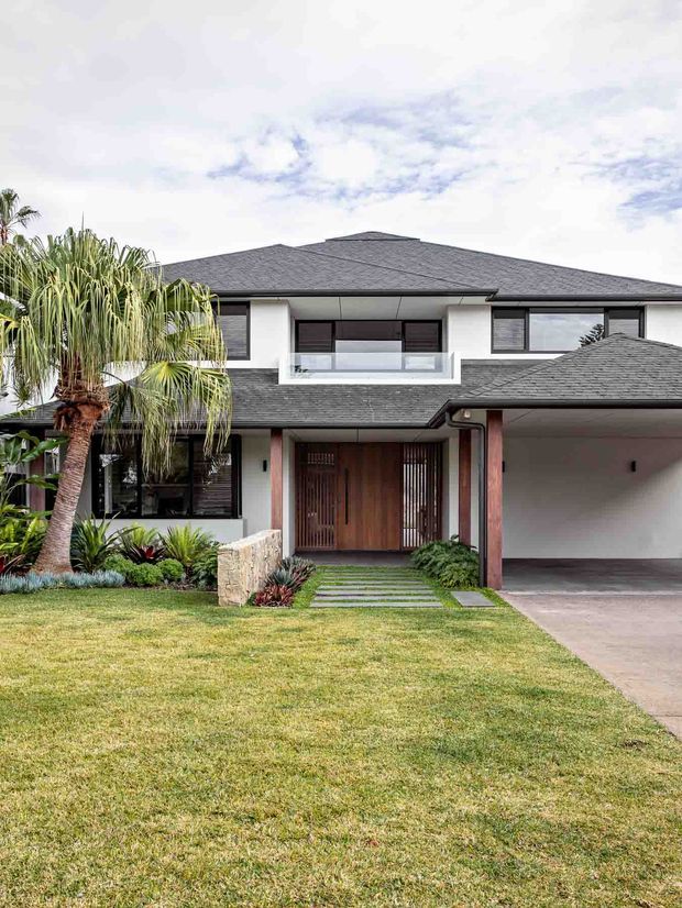 Front exterior view of a large contemporary two-storey residence with a dark hipped roof, white rendered walls, extensive dark-framed glazing, a recessed timber entry door, covered balcony, and an attached carport. Tropical landscaping, lawn, stepping stones, and a driveway frame the house.