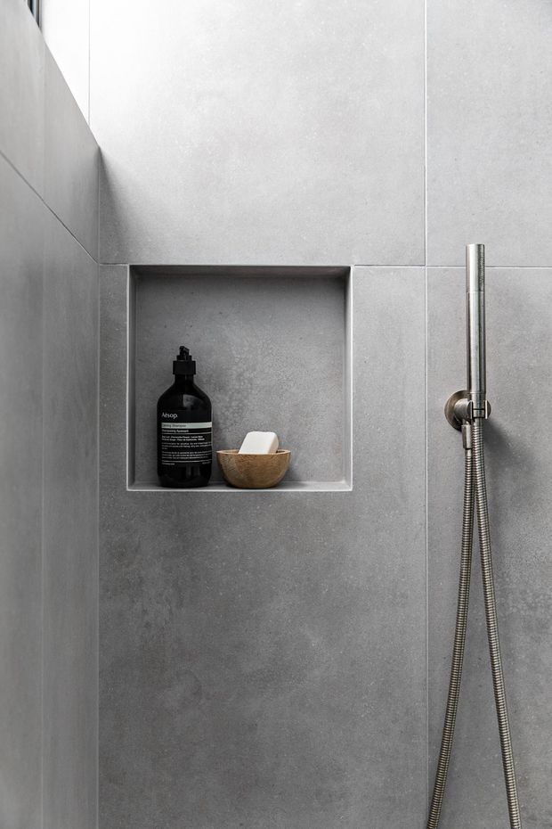 Close-up view of a minimalist shower wall clad in large-format grey tiles, with a recessed rectangular niche containing toiletries and a small timber bowl. A slim chrome handheld shower rail and flexible hose are mounted on the right.