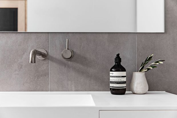 Close-up of a contemporary bathroom vanity with a white integrated basin and countertop, grey stone-look wall tiles, brushed metal wall-mounted tapware, and a minimalist mirror. A dark soap dispenser and small ceramic vessel with greenery sit on the counter.