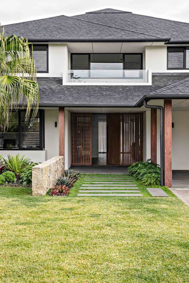 Front exterior view of a contemporary two-storey home with a broad dark shingle roof, white rendered walls, dark-framed windows, timber screening and entry doors, and a recessed covered entrance. A glass-balustraded upper balcony sits above the entry, while stone edging, layered planting and a stepping-stone path lead through the lawn.