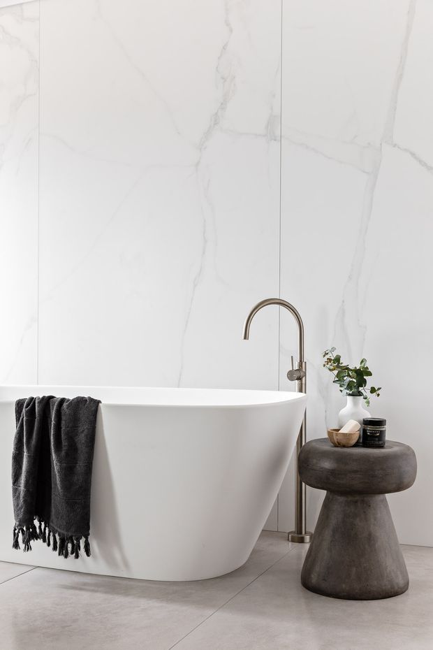 A refined contemporary bathroom featuring a freestanding white bathtub against large-format white marble-look wall panels, with a brushed metal floor-mounted tub filler. A dark charcoal towel hangs over the tub, beside a sculptural dark stone or concrete stool holding toiletries and a small plant. The floor is a light grey concrete-look finish, creating a calm, minimalist spa-like setting.