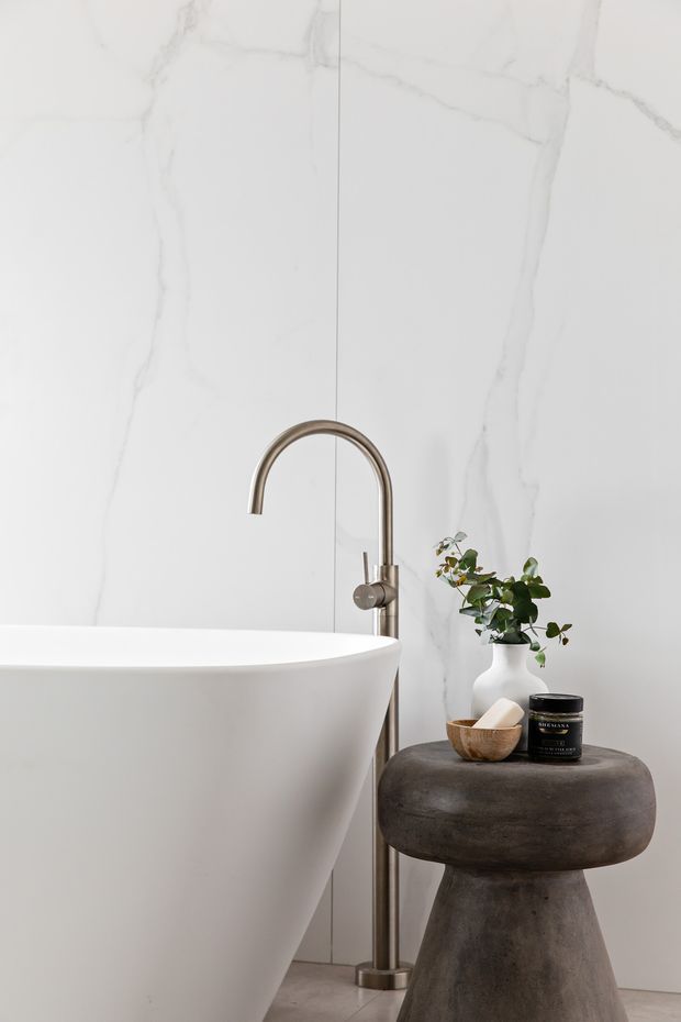 A close-up of a refined contemporary bathroom featuring a freestanding white oval bathtub, a brushed metallic floor-mounted gooseneck faucet, and large white marble-look wall slabs with subtle grey veining. A dark sculptural pedestal stool beside the tub holds a vase with greenery and small accessories.