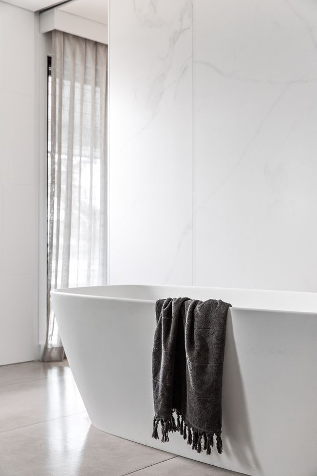 Bright minimalist bathroom featuring a freestanding white bathtub, large white marble-look wall panels, sheer floor-to-ceiling curtains, and a pale tiled floor. A dark charcoal towel drapes over the tub, providing contrast against the restrained white and grey palette.
