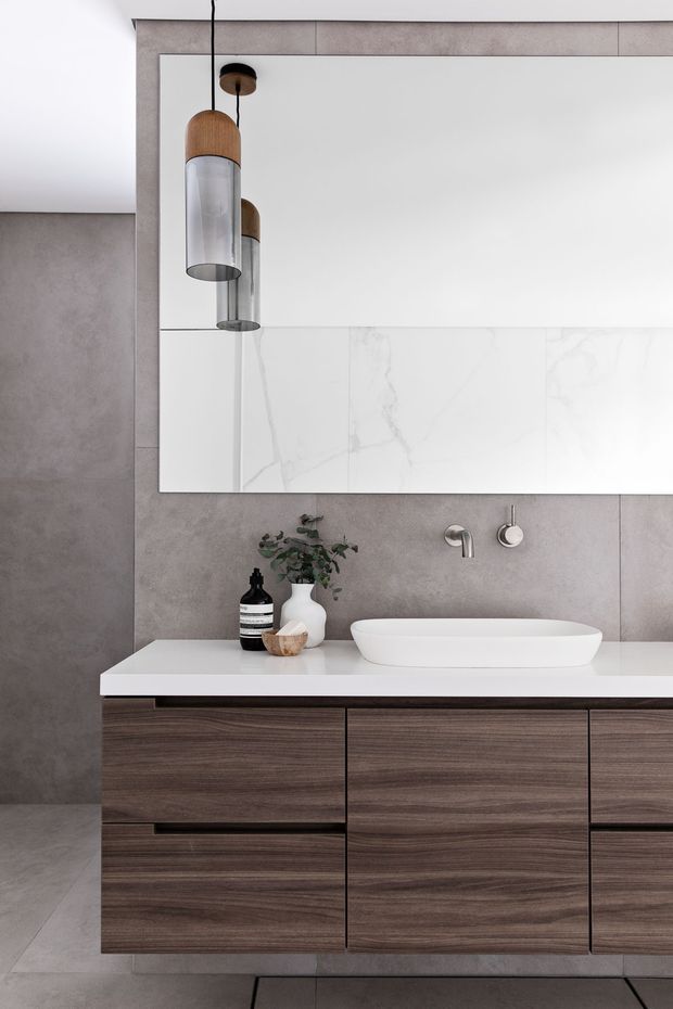 Contemporary bathroom vanity featuring a wall-mounted dark timber joinery unit, white integrated basin and benchtop, large frameless mirror, grey stone-look wall and floor tiles, and cylindrical pendant lighting with timber accents.