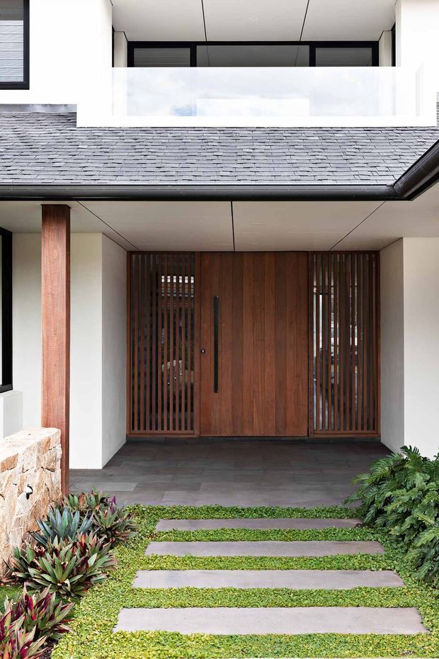 A contemporary residential entry viewed from the landscaped front path. The recessed entrance features a large warm timber door with matching vertical timber screening, dark slate-like paving, white rendered walls, a timber support post, and a covered ceiling. A grey slate-clad roof and upper-level balcony or opening are visible above, with lush low planting and stepping stones leading to the entry.