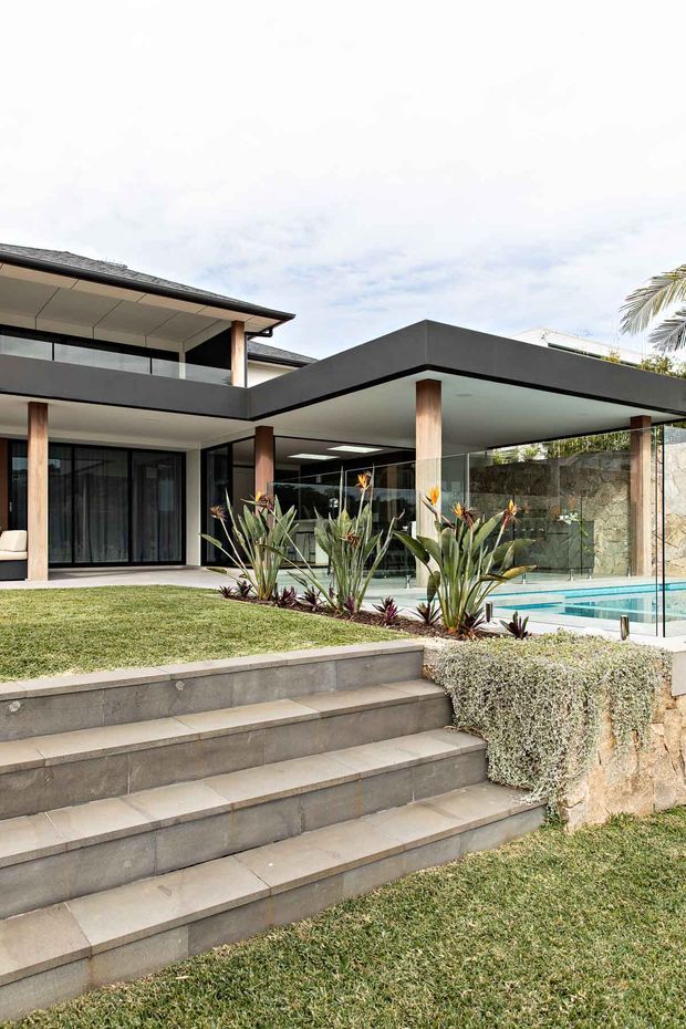 Exterior view of a contemporary multi-level residence with broad flat roof planes, dark charcoal fascia and trim, light timber-clad columns, extensive glazing, and a covered outdoor living area beside a swimming pool. Landscaped steps, lawn, clipped hedging, tropical planting, and stone retaining walls define the foreground and garden.
