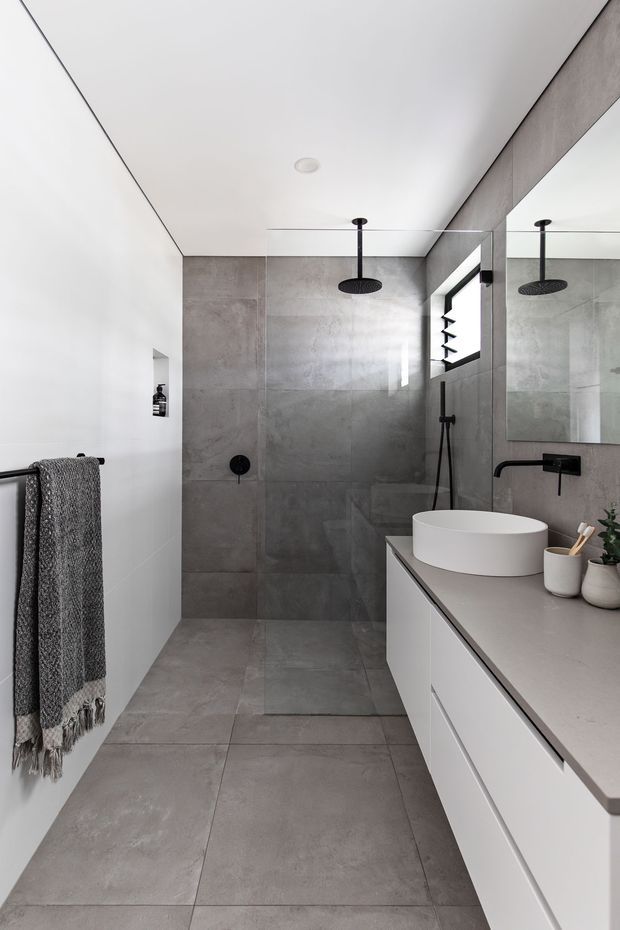 A contemporary bathroom with a walk-in shower, large-format grey concrete-look wall and floor tiles, matte black shower and tapware, a round vessel basin on a white floating vanity, and a large mirrored wall. The space is bright, restrained, and monochromatic with soft grey and white finishes.