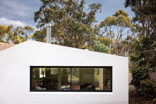 A single-storey white rendered residential building set among mature eucalyptus trees. The simple low-pitched roof is largely concealed behind a clean parapet-like facade, with a prominent wide black-framed glazed window revealing a contemporary interior. A small section of terracotta tiled roofing and a metal chimney are visible at the left.