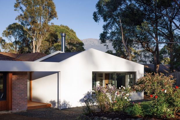 Single-storey contemporary house exterior with a crisp white rendered gabled facade, broad low-pitched roof, dark roof-mounted chimney, exposed red-brick wall, timber-framed entry, and large glazed window. Mature eucalyptus trees and planted garden surround the home, with a rugged mountain visible in the background.