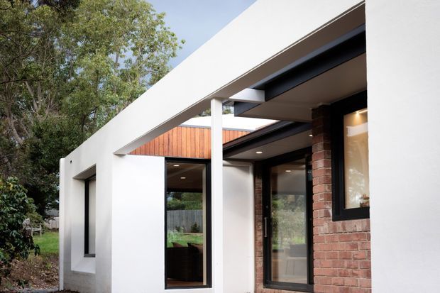 Exterior view of a contemporary single-storey home entrance, featuring crisp white rendered walls, dark-framed glazing, exposed red brickwork, timber cladding, and a projecting flat roof or canopy. Mature trees and garden landscaping surround the facade.