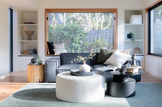 Bright, carefully styled living room with a dark charcoal sofa, sculptural white boucle ottoman, black round coffee tables, and built-in open shelving. Large timber-framed windows bring in abundant natural light and overlook greenery, while pale walls and light timber flooring create an airy, contemporary setting.