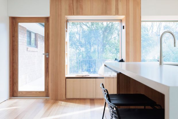 Bright contemporary kitchen interior with extensive vertical timber wall panelling and matching timber cabinetry, a white waterfall-edge island with a tall curved metal tap, and black bar stools. Large windows and a glazed timber-framed door bring in views of trees and abundant natural light. The space has a clean, warm, minimalist character.