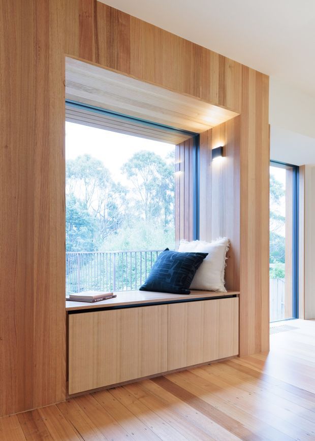 A bright, contemporary interior featuring a built-in timber window seat and storage cabinetry set within a full-height timber wall lining. The recessed nook has cushions and a dark throw pillow, with large windows opening to leafy views and a balcony railing. Warm timber flooring, integrated wall lighting, and clean geometric detailing create a calm, refined reading or sitting area.