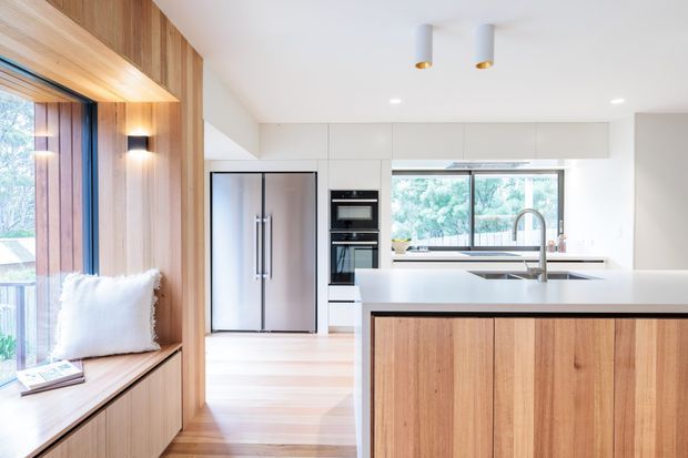 Bright contemporary kitchen with a large timber-clad island, white cabinetry and benchtops, integrated stainless-steel appliances, and a generous window overlooking greenery. A built-in timber window seat with cushions occupies the left side, while recessed and cylindrical ceiling lighting create a clean, warm interior.