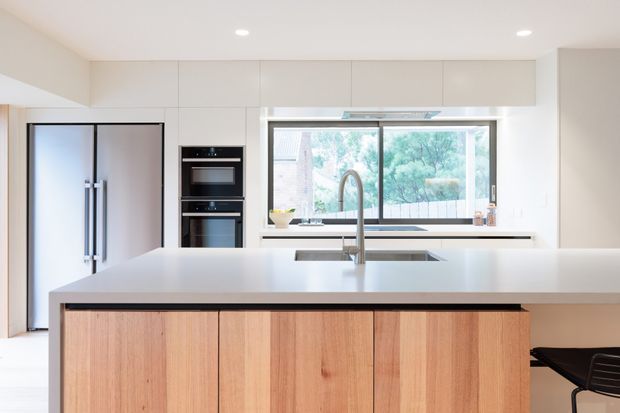 Bright contemporary kitchen viewed across a large central island. The space features white full-height cabinetry, integrated black ovens, a large stainless-steel refrigerator, a pale stone island benchtop with an undermount sink and sculptural mixer tap, and warm timber-grain island cabinetry. A wide window above the rear benchtop brings in abundant natural light and overlooks greenery. A partial black bar stool is visible at the island.