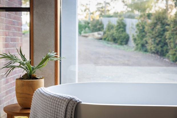 A close-up of a freestanding white bathtub positioned beside large windows. A textured grey throw hangs over the tub edge, with a potted plant on the sill or adjacent ledge. Exposed red brick, timber window framing, and a view toward a landscaped driveway create a warm, natural bathroom setting.