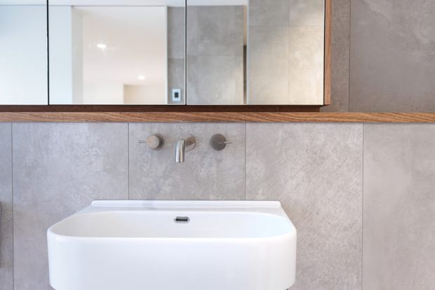 Close-up of a contemporary bathroom vanity area featuring a white freestanding basin, wall-mounted brushed metal tapware, large pale grey stone-look wall tiles, a timber-framed mirrored cabinet, and a timber shelf or ledge. The palette is soft grey, white, and warm wood with clean minimalist detailing.