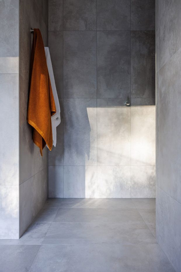 Minimalist walk-in shower or wet-room interior lined entirely with large-format cool grey concrete-look tiles. The floor and walls form a seamless, restrained enclosure with a small chrome control fitting and wall-mounted towels, including a warm burnt-orange towel that adds the only strong colour accent. Natural light creates a bright vertical patch across the rear wall and floor.