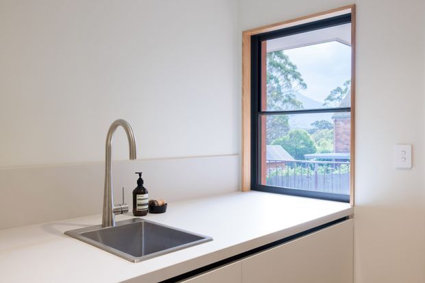 Bright, minimalist kitchen work zone featuring a white benchtop, integrated stainless-steel sink and gooseneck tap, pale fixed cabinetry, and a large black-framed timber-trimmed window overlooking greenery and neighbouring rooftops. The restrained palette and clean surfaces create a fresh, functional atmosphere.