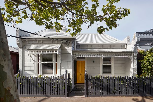 Street-facing view of a compact heritage-style single-storey residence with a pale rendered or weatherboard-like facade, corrugated metal hipped roof, decorative eaves and window awnings, timber louvred windows, and a bright yellow front door. A dark vertical picket fence and narrow entry path frame the facade, with mature street trees in the foreground. An ornate neighbouring building is partially visible at right.