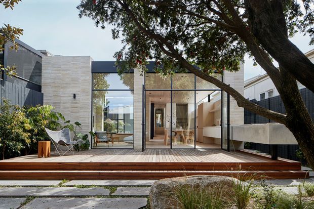 Exterior view of a contemporary house opening onto a broad timber deck and landscaped garden. The composition features a strong rectilinear form, pale masonry walls, dark cladding, extensive floor-to-ceiling glazing, and a sheltered courtyard-like outdoor living area. Interior living and dining spaces are visible through the glazing, while mature planting and a large tree frame the foreground.