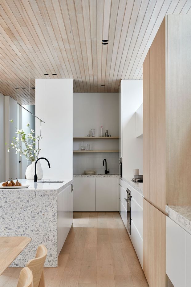 A bright, refined contemporary kitchen with a galley-style layout and a central terrazzo island. The kitchen features pale timber cabinetry, white cabinetry, integrated appliances, a light timber slatted ceiling, pale timber flooring, open shelving, a ribbed splashback, and black gooseneck tapware. A dining table and chairs are partially visible in the foreground.