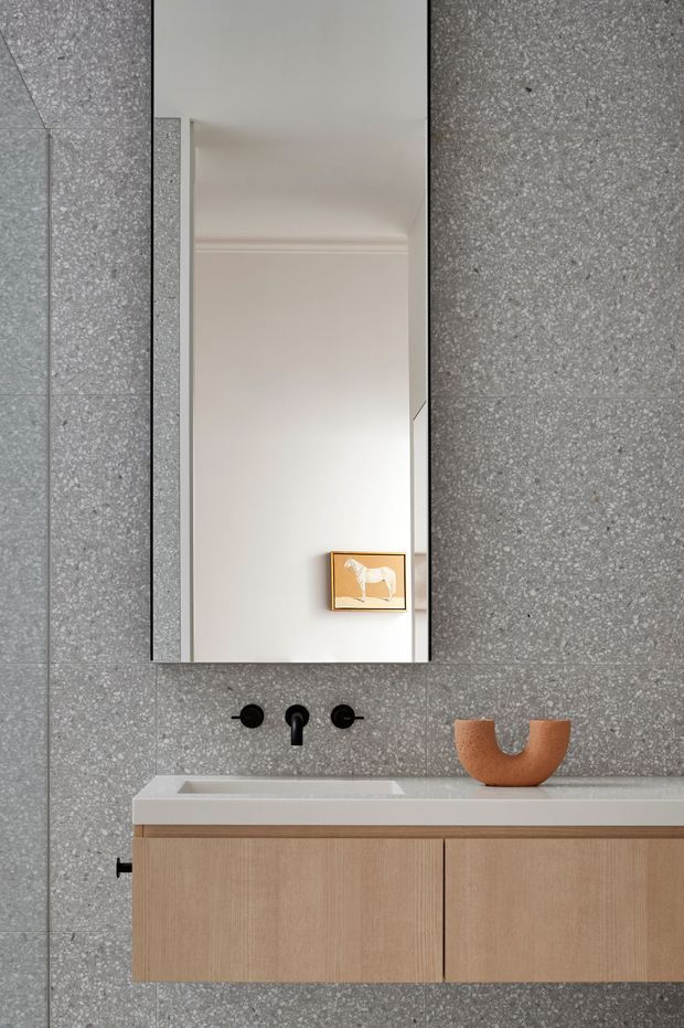 A compact, refined bathroom vanity featuring grey terrazzo-look wall and floor tiles, a floating light timber vanity with a white integrated basin, and matte black wall-mounted tapware. A tall frameless mirror reflects a softly moulded white ceiling and framed artwork, while a small terracotta sculptural vessel adds warmth.