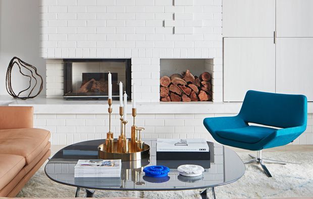 Bright contemporary living room featuring a white painted brick fireplace wall with a recessed firebox and stacked firewood niche, a tan sofa, sculptural blue armchair, and a reflective oval glass coffee table with brass candlesticks and decorative objects. The room has a clean, refined mid-century-influenced interior with strong blue and warm neutral accents.