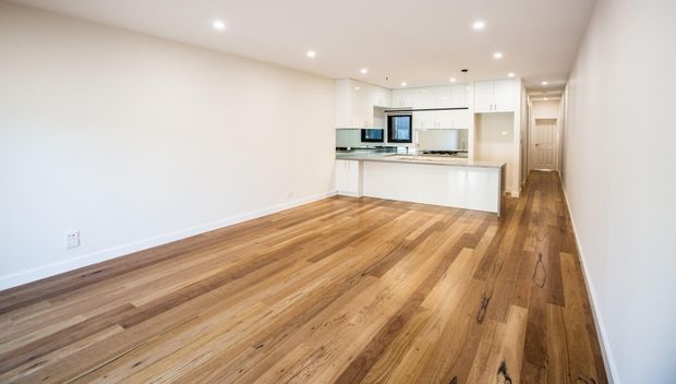 Bright, open-plan modern apartment interior with a predominantly empty living area leading into a compact white kitchen. The space features warm timber flooring, white walls and cabinetry, a stone-look island benchtop, recessed ceiling lights, and a narrow hallway extending beyond the kitchen.