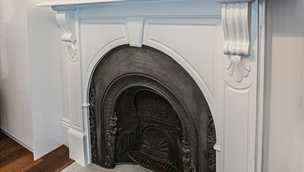 Close-up of an ornate white-painted decorative fireplace surround with a dark cast-iron arched fire insert. The surround features carved classical detailing, fluted side elements, layered mouldings and floral ornament, set against pale walls and a dark timber floor.