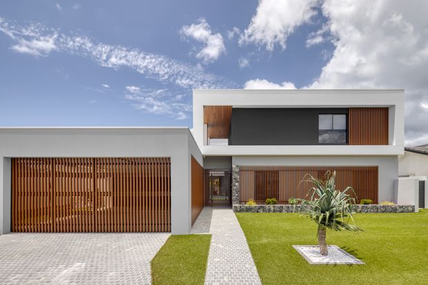 Contemporary two-storey residence with crisp white rendered walls, a flat roofline, dark recessed upper-level glazing, and extensive warm timber vertical screening. A broad timber-slatted garage door sits to the left, while a patterned paved path leads through a manicured lawn to the entry. The front garden includes a small feature palm and low stone-edged planting beds under a bright blue sky.
