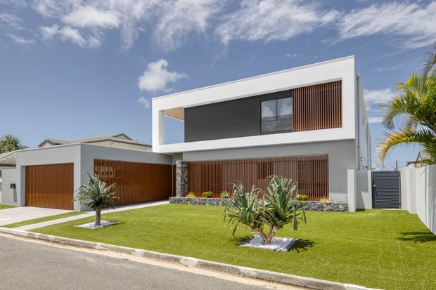 Street-facing view of a contemporary two-storey residence with crisp white rendered walls, strong rectangular geometry, broad dark-framed glazing, and extensive warm timber vertical screening across the garage and ground-floor frontage. The property has a manicured lawn, tropical planting, a paved pedestrian path, and a slatted side gate. A flat roof and clean minimalist facade dominate the composition.