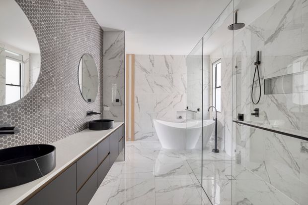 Contemporary luxury bathroom with a freestanding white bathtub, large-format white marble-look wall and floor surfaces, a frameless glass shower enclosure, and a long double vanity with black vessel basins. A curved wall section is covered in small grey mosaic tiles, complemented by black tapware, shower fittings, mirrors, and slim dark-framed windows.