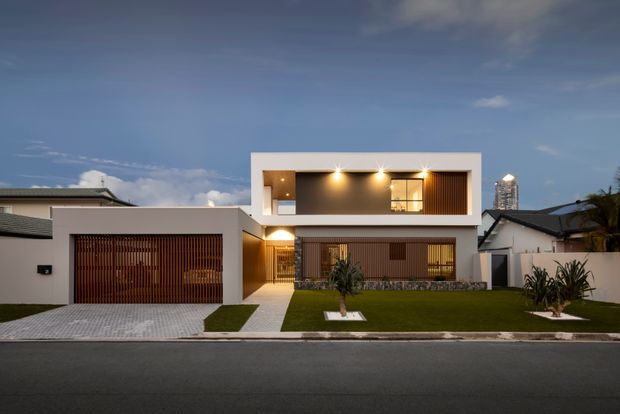 Street-facing photograph of a contemporary two-storey residence at dusk. The home has a crisp white rendered facade, a strong rectangular upper-level volume with a recessed illuminated balcony or terrace, vertical timber screening across the garage and lower windows, a stone-clad low wall, and a neatly landscaped front lawn with paved paths. A neighbouring house and distant urban high-rise are visible in the background.
