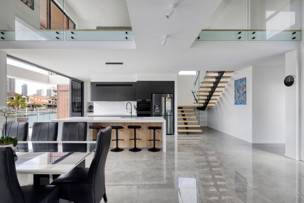 Bright, spacious open-plan interior with a contemporary kitchen, dining area and floating staircase. The kitchen features matte black cabinetry, a white island with seating, stainless-steel appliances and a reflective grey polished floor. Full-height glazing opens views toward a waterfront or urban setting, while a glass balustrade overlooks the lower level, creating a mezzanine-like double-height space.