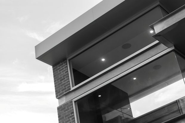 Close-up, low-angle exterior view of a contemporary multi-level building corner. Dark brickwork, large framed glazed openings, projecting box-like roof and cantilevered slab elements create a strong geometric composition, with recessed ceiling lights visible beneath the overhangs. The image is monochrome.