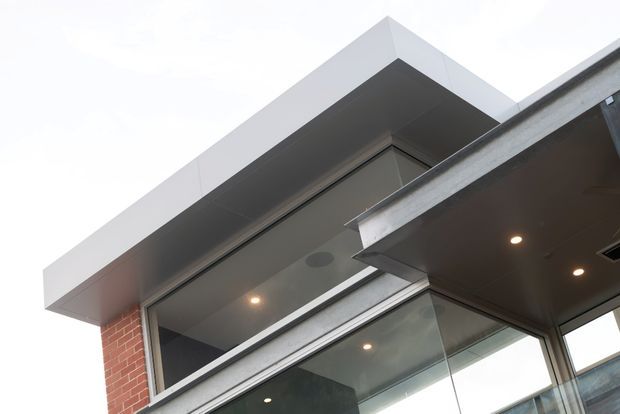 Close-up upward view of a contemporary building facade, showing a projecting box-like roof or canopy, dark grey soffits with recessed downlights, large glazed openings in aluminium frames, and a section of exposed red brick wall. The composition emphasises clean geometric lines and layered horizontal projections.