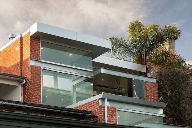 Exterior view of a multi-level contemporary brick residence with strong geometric forms, broad floor-to-ceiling glazing, projecting flat roof planes, and dark grey metal fascia and trim. A partially covered upper terrace or outdoor living area is visible, along with palms behind the building. The facade combines red brickwork, pale grey rendered or metal surfaces, and extensive glass.