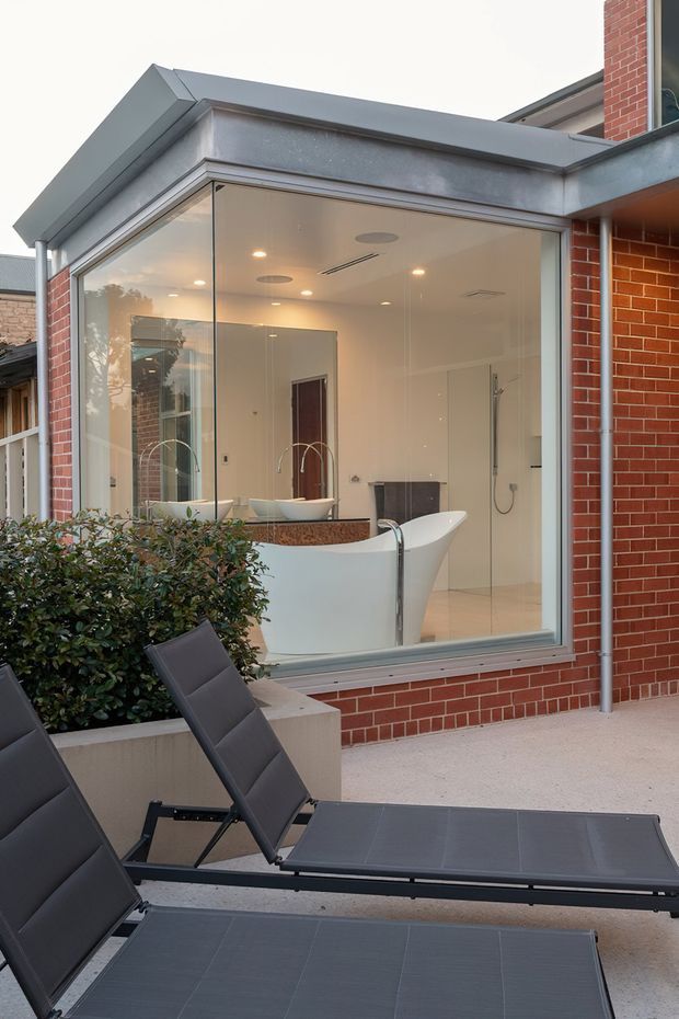 Exterior view of a contemporary brick-clad home addition with a fully glazed bathroom enclosure. The bathroom visibly contains a freestanding white bathtub, wall-mounted shower, double basins, large mirror, and warm recessed ceiling lights. In the foreground is a paved outdoor terrace with dark reclining sun loungers and raised planter boxes.