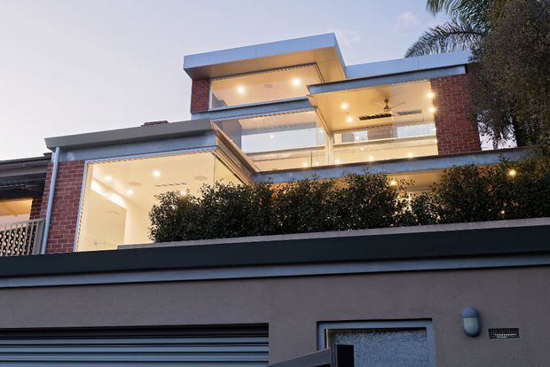 Twilight street-level view of a contemporary multi-level residential facade. The house features red brick walls, crisp white and grey horizontal roof slabs and balcony edges, extensive glazing, recessed warm lighting, covered outdoor terraces, and a high front boundary wall with dense hedging. Palm and other vegetation frame the upper right of the image.