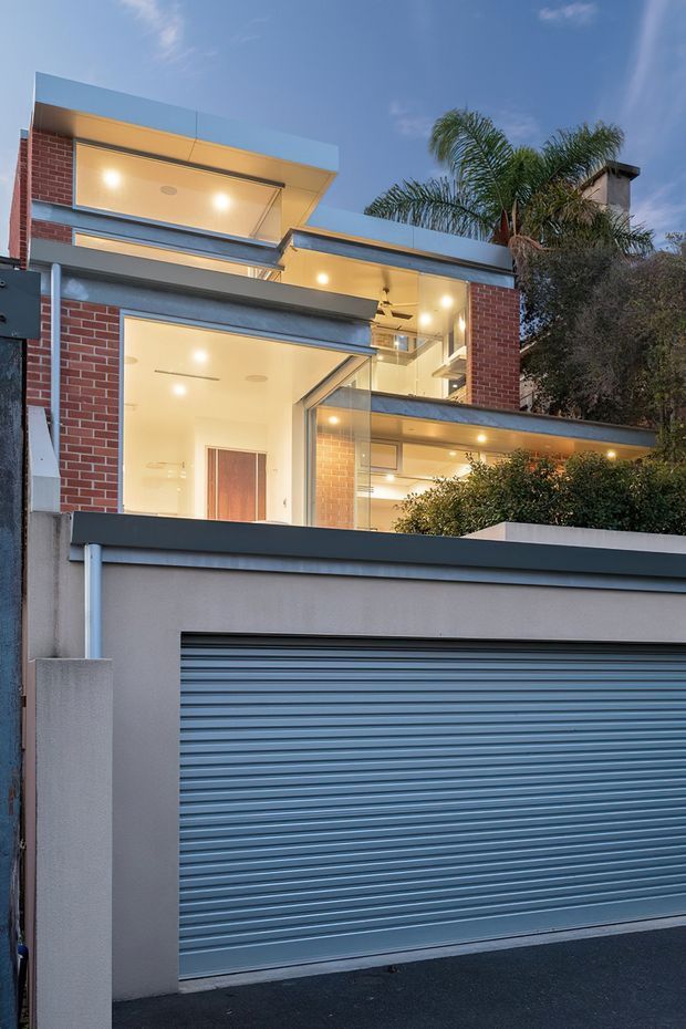 Exterior view of a contemporary multi-level urban residence at dusk, seen behind a front boundary wall and roller garage door. The facade combines red brick, smooth light render, dark metal roof and trim elements, extensive glazing, projecting flat roof forms, and warm interior lighting. Tropical palms and landscaping are visible around the house.