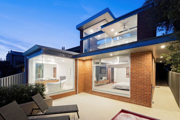 Twilight exterior view of a contemporary multi-level brick residence centred on a private paved courtyard or terrace. Large glazed openings reveal a bright bathroom on the left and a bedroom on the right, while an upper-level living area or landing is visible through expansive windows. The home has strong horizontal roof planes, projecting eaves, dark window frames, exposed red brick walls, and integrated exterior lighting.