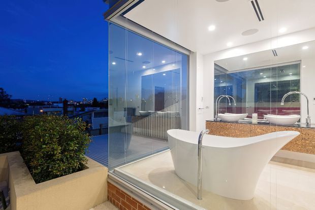 A contemporary bathroom viewed from an exterior terrace at dusk, featuring a freestanding white sculptural bathtub, dual vessel basins, wall-to-wall mirrors, a warm speckled stone vanity, white tiled surfaces, and extensive floor-to-ceiling glazing. The urban or waterfront-like view is visible beyond the glass, with a planted raised trough in the foreground.