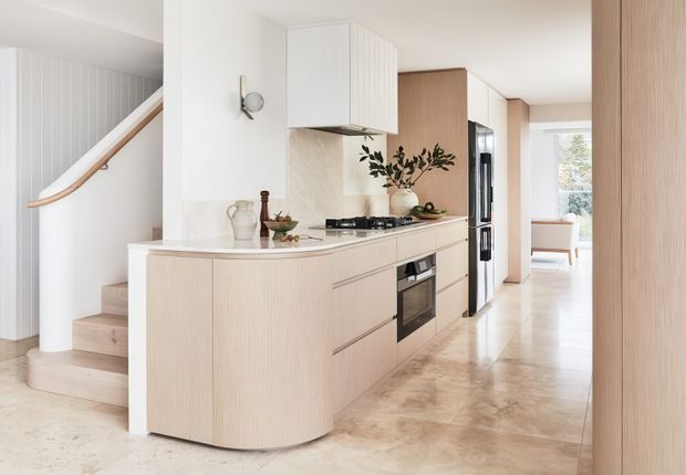 Bright contemporary kitchen with a curved light-wood island and integrated cabinetry, pale stone benchtops and splashback, a compact cooktop and built-in oven, and a ribbed white rangehood cabinet. A curved timber handrail and staircase are visible to the left, while a hallway and bright sitting area extend beyond the kitchen. The space has a refined, warm-neutral palette and clean built-in detailing.